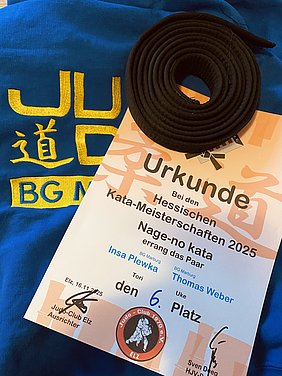 Das Bild zeigt ein blaues T-Shirt mit gelbem Text, einen schwarzen Judogurtel und eine Urkunde. Die Urkunde bestätigt den 6. Platz bei den Hessischen Kata-Meisterschaften 2025 für das Paar Insa Plewka und Thomas Weber in der Kategorie Nage-no kata.