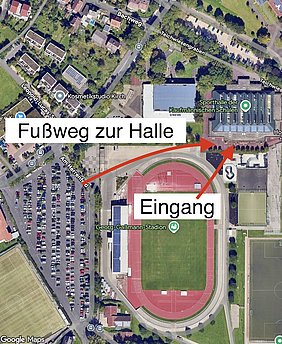 Eine Übersichtskarte zeigt den Weg zur Halle. Ein Fußweg führt vom Georg-Gaßmann-Stadion zur Sporthalle der Kaufmännischen Schulen. Der Eingang ist klar markiert. Die Umgebung umfasst parkende Autos und angrenzende Gebäude.