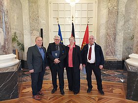 Vier Personen stehen in einem großen Raum mit marmornen Wänden. Im Hintergrund sind Flaggen sichtbar. Die Gruppe sieht freundlich aus; eine Person hält eine Mappe. Die Atmosphäre wirkt feierlich und offiziell.