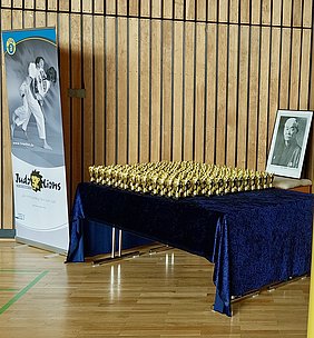 In einem Hallenraum stehen auf einem Tisch zahlreiche goldene Pokale in Reihe. Neben dem Tisch befindet sich ein gerahmtes Porträt eines älteren Mannes. Ein Banner mit dem Logo „Judo Lions“ steht ebenfalls in der Nähe.