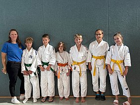 Sechs Kinder in weißen Judokis mit verschiedenen farbigen Gürteln stehen in einer Reihe. Links steht eine Frau in einem blauen T-Shirt. Hintergrund ist eine graue Wand. Die Kinder lächeln und posieren freundlich für das Foto.