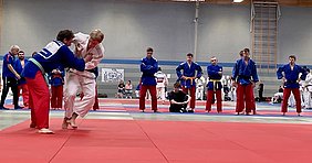 Zwei Judoka kämpfen auf einer Matte. Einer trägt einen weißen Gi und wird von einem Judoka in blauer Kleidung gegriffen. Im Hintergrund stehen mehrere Zuschauer und weitere Judoka, die das Geschehen beobachten. Die Atmosphäre ist競争lich und konzentriert.