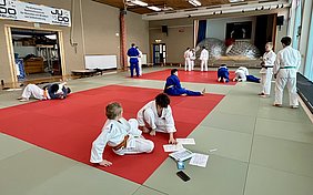 Eine Gruppe von Judokas trainiert auf einer Matte, die teilweise rot und teilweise grün ist. Einige üben Techniken, während andere in einer Sitzung sitzen oder auf Notizen schauen. Im Hintergrund ist eine Bühne mit einer großen Wand und einem Banner zu sehen.