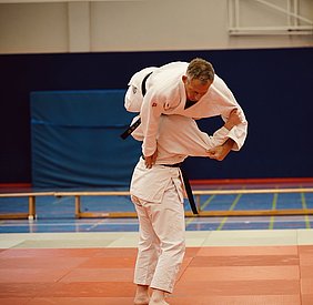 Zwei Judokas in einem Trainingsraum. Ein Kämpfer wird von einem anderen Spieler im Wurf gehalten. Die Athleten tragen traditionelle Judo-Gewänder in Weiß und Schwarz. Der Boden ist mit Judomatten in verschiedenen Farben ausgelegt.