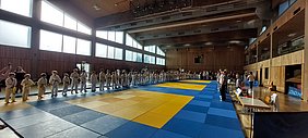 Eine Gruppe von Kindern in weißen Judogi steht in zwei Reihen auf einer Judomatte. Einige tragen farbige Gürtel. Im Hintergrund sind Zuschauer zu sehen, die die Veranstaltung beobachten. Der Raum hat große Fenster, durch die Tageslicht einfällt.