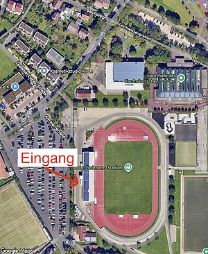 Die Karte zeigt den Eingang zum Georg-Gaßmann-Stadion, markiert mit einem Pfeil. In der Umgebung sind Parkplätze und benachbarte Gebäude, darunter eine Sporthalle. Die Anordnung der Straßen und Parkplätze ist ebenfalls sichtbar.