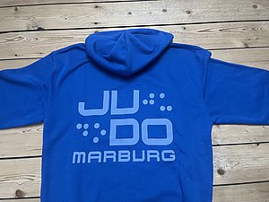 Ein blauer Kapuzenpulli mit dem Text "JU DO MARBURG" in großen, auffälligen Buchstaben auf der Rückseite. Der Schriftzug ist in einer helleren Farbe gehalten und hebt sich deutlich vom Stoff ab.