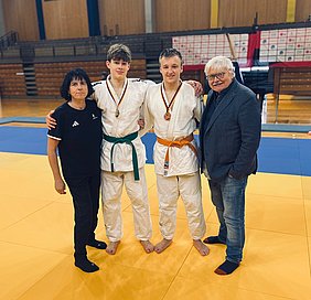 Drei Personen stehen auf einer Judomatte: zwei Jugendliche in weißen Judogi mit Medaillen, einer trägt einen grünen, der andere einen orangenen Gürtel. Neben ihnen steht eine Trainerin und ein älterer Mann. Alle wirken stolz und lächeln.