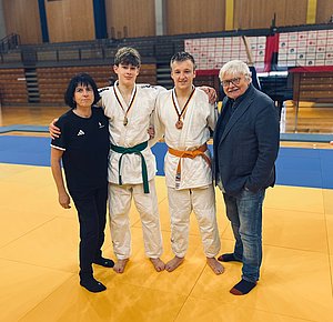 Drei Personen stehen auf einer Judomatte: zwei Jugendliche in weißen Judogi mit Medaillen, einer trägt einen grünen, der andere einen orangenen Gürtel. Neben ihnen steht eine Trainerin und ein älterer Mann. Alle wirken stolz und lächeln.