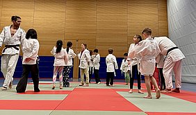 Eine Gruppe von Kindern in weißen Judokis steht auf einer Matte mit roten und grünen Bereichen. Ein Erwachsener mit braunem Gürtel zeigt eine Technik. Die Kinder sind aufmerksam und scheinen zu üben oder zu lernen. Die Atmosphäre wirkt konzentriert und sportlich.