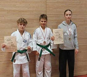 Drei Kinder in Judogis mit grünen Gürteln stehen nebeneinander. Sie halten Medaillen und Urkunden in den Händen, die ihre Erfolge dokumentieren. Die Jungen tragen jeweils eine Medaille um den Hals, während das Mädchen ebenfalls für ihre Leistungen ausgezeichnet wurde.