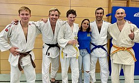 Fünf Personen in Judo-Anzügen stehen zusammen und lächeln. Ein junger Mann in der Mitte hält einen Pokal. Die Gruppe zeigt Stolz und Freude, während sie vor einer holzvertäfelten Wand posieren. Zwei Personen tragen blaue und zwei weitere orangefarbene Gürtel.