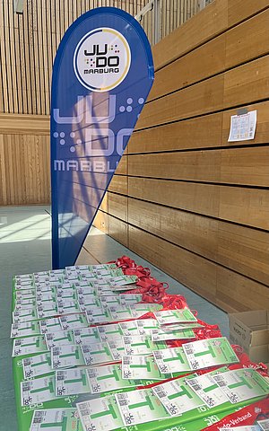 Im Bild sind ein Banner mit dem Logo des Judovereins Marburg und ein Tisch mit vielen Namensschildern zu sehen. Die Schilder sind auf grünem Karton mit roten Bändern befestigt. Der Hintergrund zeigt eine Holzwand in einer Sporthalle.