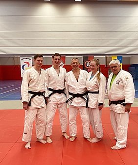 Eine Gruppe von fünf Judoka steht auf einer Matte in einem Dojo. Alle tragen weiße Judoanzüge mit schwarzen Gürtel. Sie lächeln in die Kamera, während im Hintergrund Judomatten und einige Flaggen sichtbar sind.