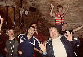Eine Gruppe von sechs fröhlichen Menschen feiert in einem gemütlichen Raum. Einige tanzen und lachen, während einer von ihnen auf den Schultern eines anderen steht. Die Atmosphäre ist lebhaft und festlich, was auf eine fröhliche Zusammenkunft hindeutet.
