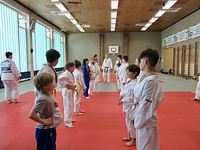 In einer Judohalle stehen Kinder in weißen und blauen Judogis in zwei Reihen. Einige tragen gelbe Gürtel. Ein Trainer mit einem schwarzen Gürtel gibt Anweisungen. Der Boden ist mit rotem Mattenmaterial ausgelegt, während Fenster für Tageslicht sorgen.