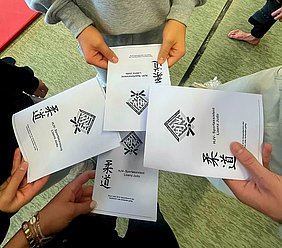 Fünf Hände halten zusammen mehrere Faltblätter mit dem Titel "HJV-Sportassistent Lizenz Judo". Auf den Blättern sind zudem japanische Schriftzeichen und ein Logo zu sehen, die auf den Inhalt und das Thema Judo hinweisen.