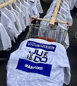 Ein Wäschekorb mit weißen Judo-Anzügen hängt an einer Stange. Einer der Anzüge zeigt das Logo "Leistungszentrum Judo Marburg" in blauer Schrift auf weißem Hintergrund.
