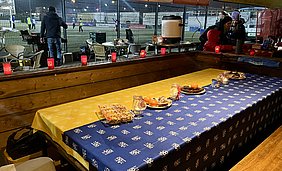 Ein gedeckter Tisch mit gelbem und blauem Tischtuch, auf dem sich verschiedene Snacks und Getränke befinden. Im Hintergrund sind Menschen, die Fußball spielen. Rote Teelichter sorgen für eine gemütliche Atmosphäre.