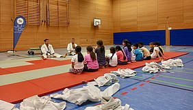 Zwei Judo-Trainer sitzen auf einer Matte und unterrichten eine Gruppe von Kindern, die in mehreren Reihen davor sitzen. Die Kinder sind aufmerksam und tragen sportliche Kleidung. Die Umgebung ist eine Sporthalle mit Holz- und Farbwänden.