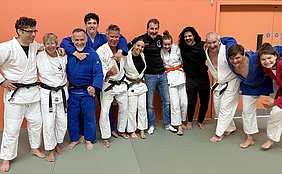 Eine Gruppe von zehn Personen steht zusammen auf einer Matte in einem Sportraum mit orangefarbener Wand. Sie tragen verschiedene Judokimonos in den Farben Weiß, Blau und Schwarz. Alle lächeln und posieren fröhlich für das Foto.