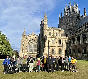 Eine Gruppe von etwa 25 Personen steht auf einer Wiese vor einer beeindruckenden Kathedrale mit hohen Türmen und großen Fenstern. Der Himmel ist blau mit vereinzelten Wolken. Einige Personen posieren und lächeln, während andere sich entspannt in die Pose setzen.