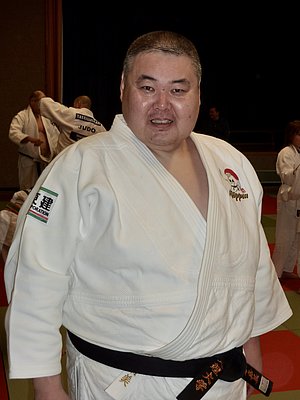 Ein männlicher Judoka in einem weißen Judoanzug und einem schwarzen Gürtel steht lächelnd im Vordergrund. Im Hintergrund sind weitere Personen in Judoanzügen während des Trainings zu sehen. Die Szenerie vermittelt eine sportliche und freundliche Atmosphäre.