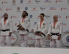 Vier Judokas stehen auf dem Siegerpodest. Der Athlet in der Mitte trägt eine Goldmedaille, während die anderen Silber- und Bronzemedaillen tragen. Alle Teilnehmer halten Blumen und tragen traditionelle Judokleidung. Der Hintergrund zeigt Sponsorenlogos und den Namen der Veranstaltung.