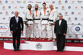 Fünf Judokas stehen auf dem Treppchen, zwei tragen Gold- und Silbermedaillen. Sie sind in weißen Judokas gekleidet. Links und rechts stehen zwei Männer in Anzügen. Der Hintergrund zeigt Logos von Sponsoren und eine Kulisse für die Veranstaltung.