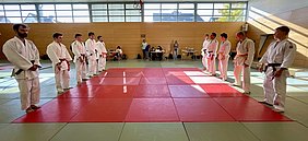 Eine Gruppe von Judokas in weißen Anzügen steht auf einer Tatami. Einige tragen rote oder schwarze Gürtel, während andere gelbe Gürtel tragen. Sie sind in zwei Reihen gegenübergestellt, bereit für einen Wettkampf oder ein Training. Im Hintergrund sind Zuschauer zu sehen.