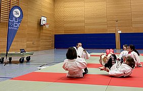 Eine Gruppe von Kindern in Judoanzügen übt auf einer Mattenfläche. Sie liegen auf dem Rücken, einige heben die Beine an. Im Hintergrund sieht man ein Basketballkorb und eine Uhr auf einem Wagen. Der Raum hat eine Holzwand und ist gut beleuchtet.