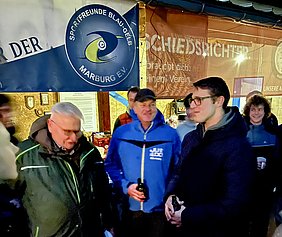 Eine Gruppe von Menschen steht zusammen an einer Veranstaltung. Im Hintergrund sind Banner der Sportfreunde Blau-Gelb Marburg e.V. und ein weiterer Hinweis sichtbar. Die Personen scheinen sich zu unterhalten und sind in Freizeitkleidung. Einige halten Getränke in der Hand.