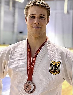 Ein junger Mann in einem weißen Judoanzug präsentiert stolz eine Bronzemedaille um seinen Hals. Auf seinem Anzug ist das Wappen Deutschlands abgebildet. Er lächelt und steht in einer sportlichen Umgebung.