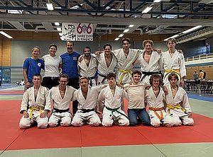 Eine Gruppe von Judokas in weißen Judogis posiert auf einer Matte. In der Mitte stehen fünf Personen, darunter auch ein Trainer. Einige tragen farbige Gürtel. Im Hintergrund sind weitere Judoka zu sehen, die an einem Training teilnehmen. Ein Banner hängt an der Decke, das das 66-jährige Bestehen des Judo in Rimbach feiert.