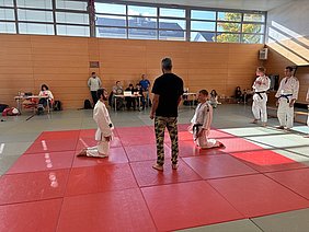 Eine Judo-Trainingssituation in einem Vereinsraum. Mehrere Kämpfer in Judogis stehen und knien auf einer roten Matte. Ein Trainer gibt Anweisungen. Im Hintergrund sind Zuschauer an Tischen zu sehen, die das Training beobachten. Tageslicht strömt durch große Fenster.