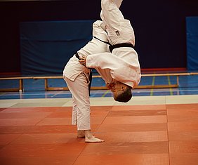 Zwei Judoka in einem Wettkampf auf einer Matte. Eine Person führt einen Wurf aus, indem sie ihren Gegner über sich hebt. Beide sind im traditionellen Judo-Gewand und tragen schwarze Gürtel. Der Hintergrund zeigt eine Sporthalle mit weiteren Matten und Bänken.