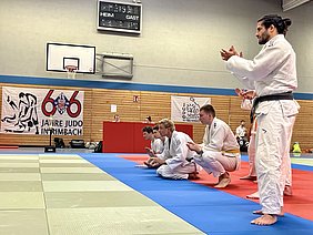 Eine Gruppe von Judokas, in weißen und blauen Anzügen, applaudiert auf der Matte. Im Hintergrund befindet sich ein Banner mit der Aufschrift "66 Jahre Judo in Rimbach". Die Judokas scheinen einen Wettkampf oder eine Vorführung zu beobachten und zeigen Unterstützung.