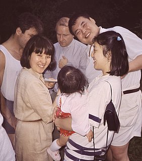 Eine Gruppe von fünf Personen, darunter zwei Frauen, die ein Baby in einem bunten Kleid halten. Alle lächeln und sehen fröhlich aus. Die Szene strahlt eine freundliche und entspannte Atmosphäre aus, umgeben von natürlichem Licht und einer grünen Umgebung.