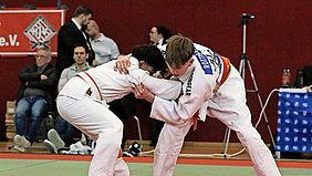 Zwei Judokas in weißen Kimonos kämpfen ineinander. Der eine Judoka hält den anderen mit beiden Händen fest. Im Hintergrund sind Zuschauer und eine rote Wand sichtbar, die eine Sportveranstaltung signalisiert.