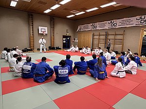 Eine Gruppe von Judoka sitzt auf der Matte in einem Trainingsraum. Ein Trainer in weißem Judogi steht vor ihnen und hält eine Ansprache. Die Teilnehmer tragen unterschiedliche Judokleidung, überwiegend in blau und weiß. Im Hintergrund sind Matten und Sportgeräte sichtbar.