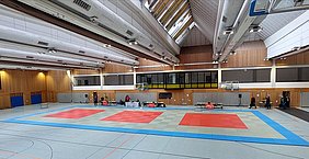 In einer Sporthalle sind mehrere Trainingsmatten in verschiedenen Farben ausgelegt. Die Matten sind in rechteckige, rote und graue Felder unterteilt. Im Hintergrund sind Tische und Personen sichtbar, die sich um Aktivitäten kümmern. Die Halle hat große Fenster und eine hohe Decke.