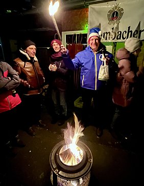 Eine Gruppe von Menschen steht in der Nacht um ein Feuer herum. Im Vordergrund brennt eine feuerspeiende Feuerstelle. Ein Mann in blauer Jacke und Nikolausmütze hält eine Fackel in der Hand und lächelt. Es herrscht eine festliche Stimmung.