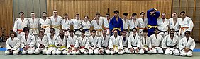 Gruppenfoto von Judo-Praktizierenden, die in weißen und farbigen Judogi (Judoanzügen) gekleidet sind. Die Athleten stehen und sitzen in mehreren Reihen, mit einigen Personen in gelben und blauen Gürteln. Der Hintergrund besteht aus Holzpaneelen und einer Judo-Matte.