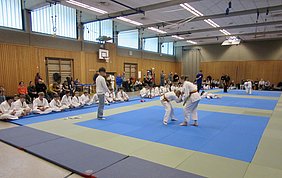 In einer Sporthalle trainieren Kinder und Jugendliche Judo. Im Vordergrund sind zwei Judoka im Wettkampf. Im Hintergrund sitzen Zuschauer und weitere Judoka, die auf Matten sitzen oder stehen. Die Atmosphäre ist konzentriert und sportlich.