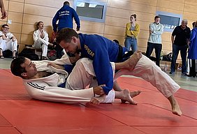 Zwei Judokas kämpfen auf einer roten Matte. Ein Kämpfer trägt einen blauen Judoanzug und versucht, seinen Gegner, der einen weißen Anzug trägt, zu kontrollieren. Im Hintergrund sind weitere Personen zu sehen, die das Training beobachten.