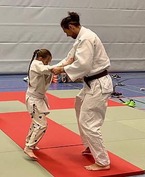 Ein Trainer und ein kleines Mädchen üben gemeinsam Judo auf einer Matte. Der Trainer zeigt dem Mädchen eine Technik, während sie ihm gegenübersteht und bereit ist, die Anweisungen umzusetzen. Beide tragen traditionelle Judokleidung.