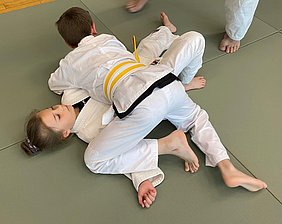 Zwei Kinder in judogerechter Kleidung kämpfen auf einer Matte. Ein Kind liegt auf dem anderen und versucht, es zu kontrollieren. Beide konzentrieren sich auf den Wettkampf, während im Hintergrund noch weitere Personen sichtbar sind.