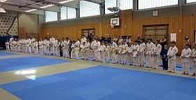 Eine Gruppe von Kindern und Jugendlichen in weißen Judogis steht auf einer blauen Matte in einem Sportraum. Die meisten haben gelbe oder orangefarbene Gürtel. Im Hintergrund sind einige Zuschauer zu sehen. Die Atmosphäre ist konzentriert und erwartungsvoll.