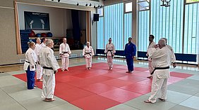 Eine Gruppe von neun Judokas in verschiedenen Judogis steht in einem Trainingsraum auf einem roten Mattenfeld. Sie scheinen sich auf eine Übung oder Demonstration vorzubereiten. Im Hintergrund hängt ein Banner mit der Aufschrift „RESPEKT erweisen“.