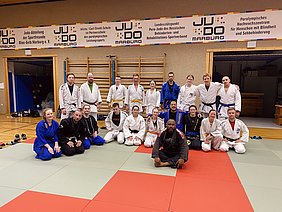 Eine Gruppe von Judokas in verschiedenen Judogis steht auf einer Judomatte. Sie sind in zwei Reihen angeordnet, einige sitzen, andere stehen. Die Umgebung ist eine Sporthalle, mit einem Banner über dem Team und dem Judosport. Alle Teilnehmer wirken freundlich und konzentriert.